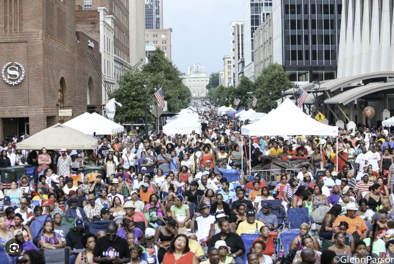A large Black community gathered at a daytime outdoor festival with tents and a city backdrop